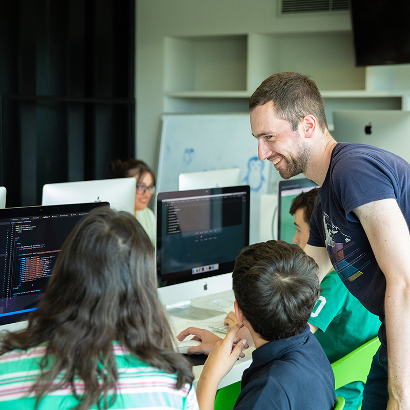 Mehrere Jugendliche vor Computern, ein erwachsener lächelt während er etwas erklärt
